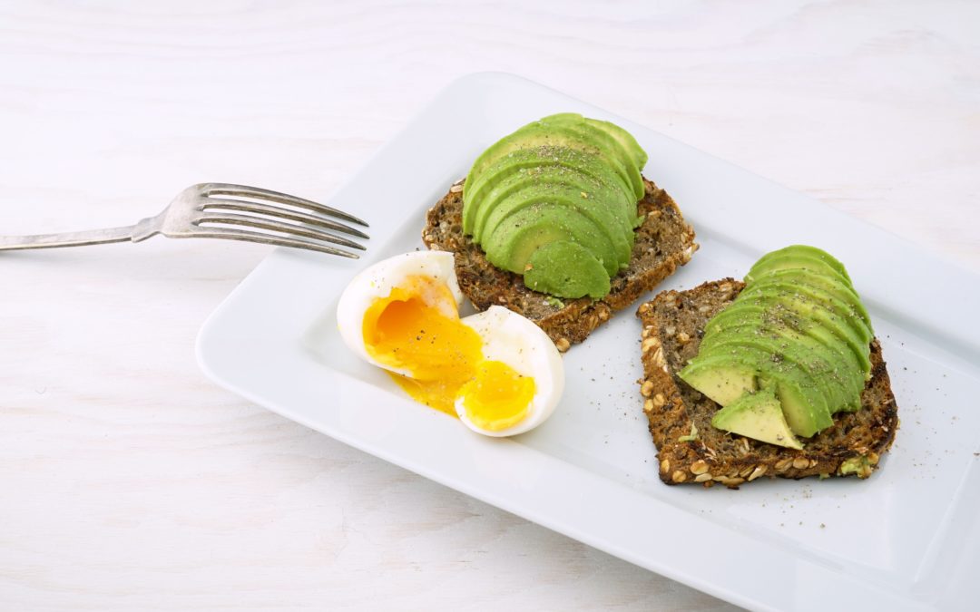 Whole wheat avocado toast on a plate with a soft-boiled egg cut in half. Avocados are a source of polyunsaturated fats which are part of a heart-healthy diet